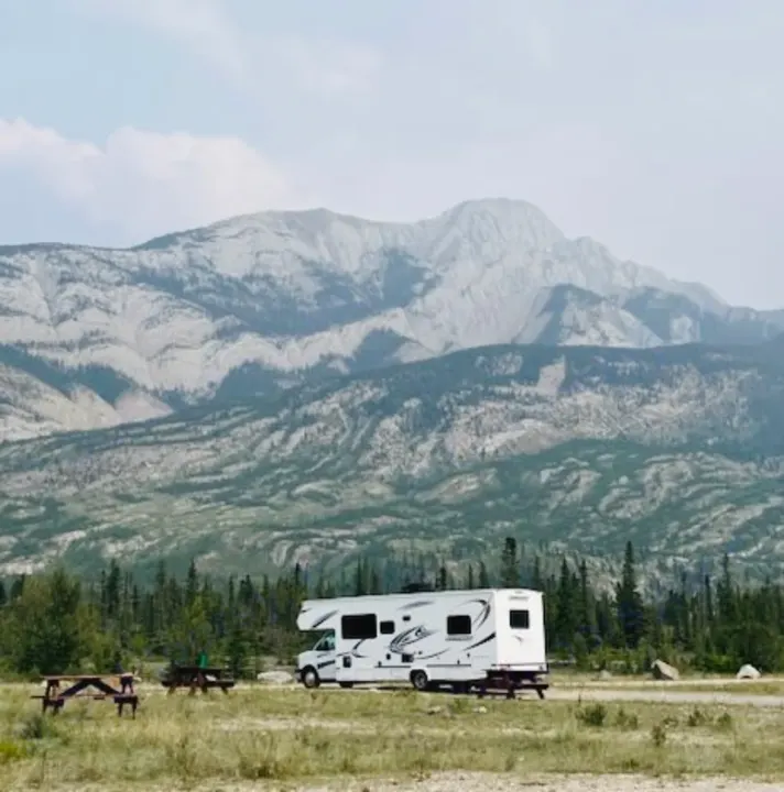 A white RV rental parked on grass with picnic tables, framed by evergreen forests and vast gray mountains.. Gulf Stream Conquest 2020