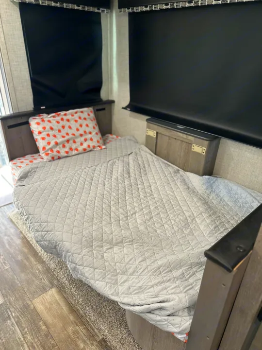 Cozy RV rental bed with a grey quilted blanket and a white, red-strawberry patterned pillow, ideal for relaxing.. Keystone RV Outback 2019