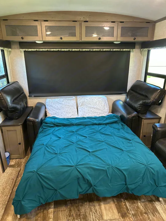 Cozy RV rental bedroom with teal bed, black leather recliners, and light wood cabinets, ideal for travel.. Keystone RV Outback 2019