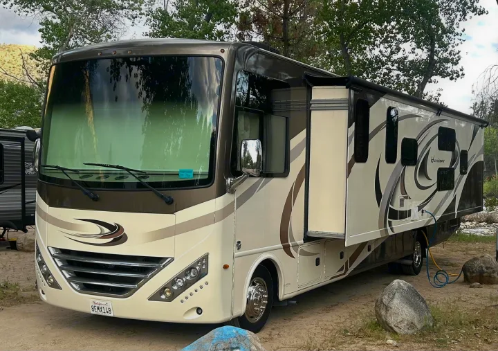 This large Class A motorhome RV rental, in beige & brown, is parked riverside with its slide-out extended for extra space.. Thor Hurricane 2018
