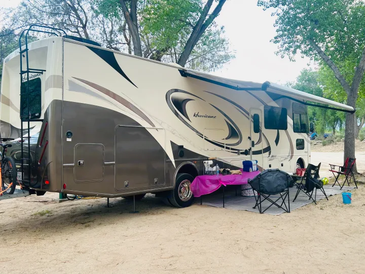Enjoy a scenic escape! This spacious RV rental is parked on sandy ground with an extended awning, chairs, and a table set up.. Thor Hurricane 2018