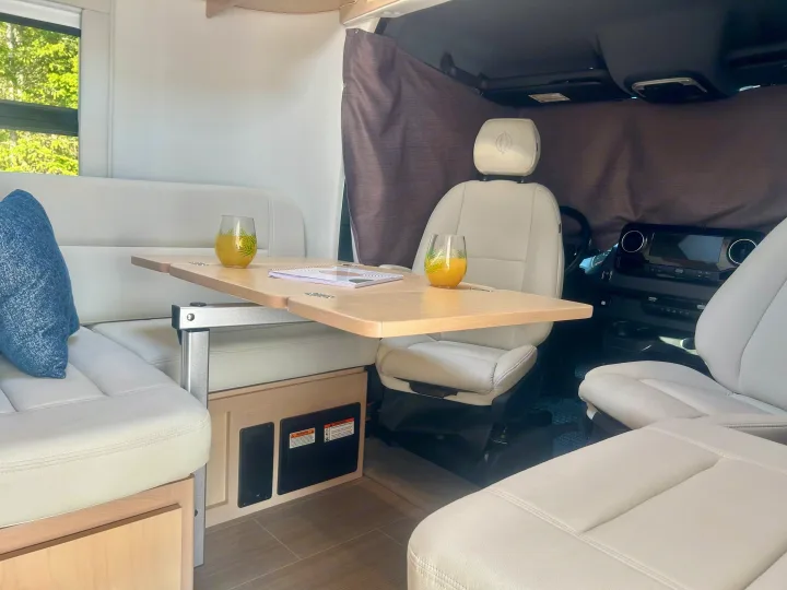 Bright RV rental interior features a dining table for two, cream lounge seating, and swivel front seats. Ideal for a camper getaway.. Leisure Travel Vans Serenity 2022