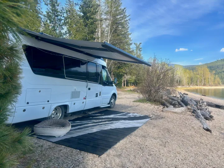 A white RV rental parked by a lake, awning extended over a mat and pet bed on the gravelly shore, surrounded by trees.. Leisure Travel Vans Serenity 2022