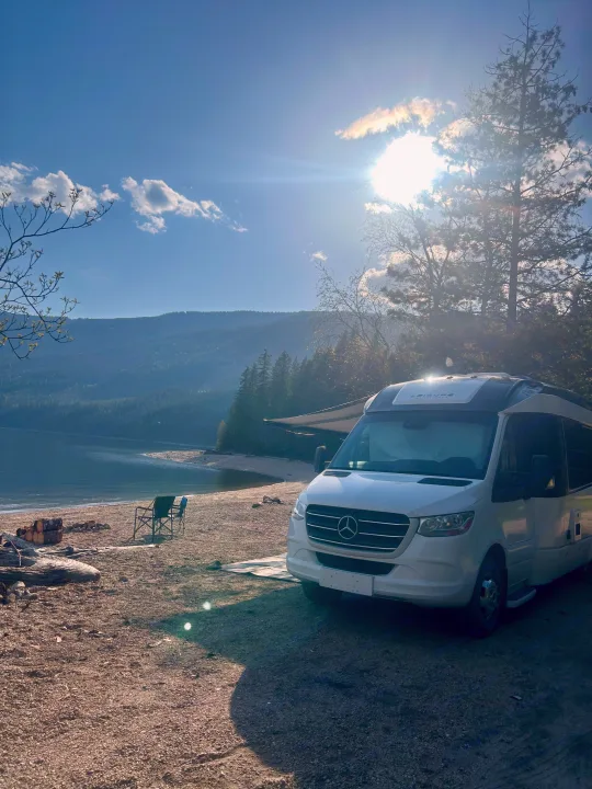 White RV rental on a serene beach by a lake, with mountains, trees, and camping chairs under a bright, sunny sky.. Leisure Travel Vans Serenity 2022