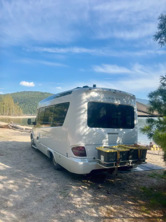 A white Leisure Travel Vans RV rental, equipped with a rear cargo carrier, is parked lakeside for scenic camping.. Leisure Travel Vans Serenity 2022