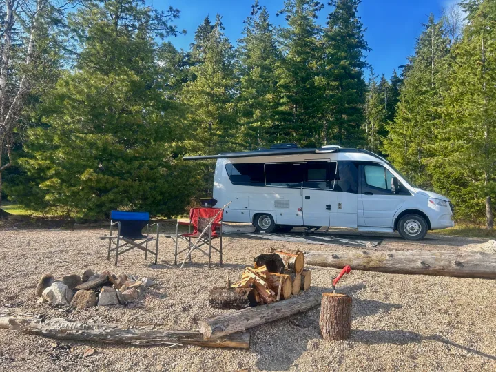 Perfect RV rental campsite setup! White camper van, fire pit, stacked firewood, and chairs among tall pines.. Leisure Travel Vans Serenity 2022