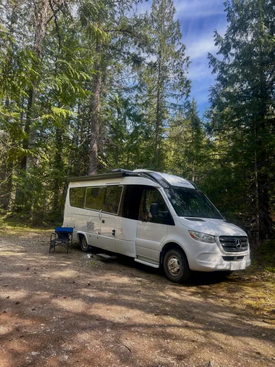 White Mercedes rv rental parked in a sun-dappled forest, perfect for a peaceful camping trip surrounded by nature.. Leisure Travel Vans Serenity 2022