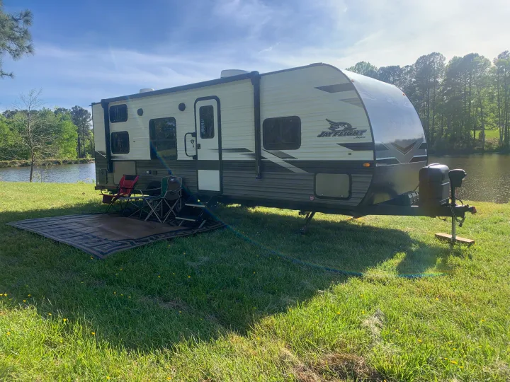 Jayco RV rental parked on a sunny lakeside spot, complete with outdoor mat and chairs for relaxing camping.. Jayco Jay Flight 2023
