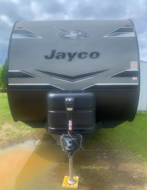 Front view of a grey and black Jayco travel trailer, ideal for an **rv rental**, with its hitch and propane tanks visible in front of a muddy puddle.. Jayco Jay Flight 2023