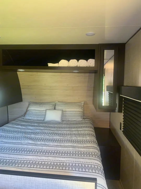 Cozy **rv rental** bedroom: patterned bed, light wood paneling, overhead storage with towels, and a mirrored cabinet.. Jayco Jay Flight 2023