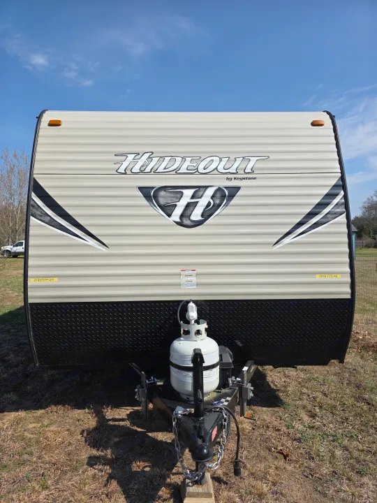 A beige Hideout by Keystone travel trailer, equipped with a propane tank and hitch, ready for an RV rental under a clear sky.. Keystone RV Hideout LHS Mini 2017