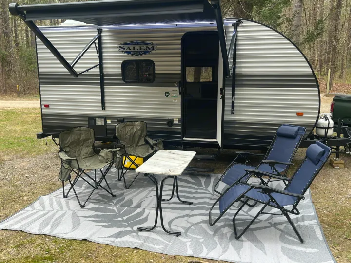 Cozy RV rental campsite: A travel trailer with extended awning, outdoor rug, green and blue chairs, and a small table.. Forest River Salem 2024