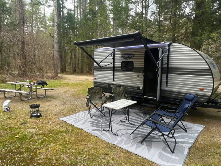 An rv rental set up at a wooded campsite, showing the travel trailer with its awning, outdoor rug, chairs, table, and grill.. Forest River Salem 2024