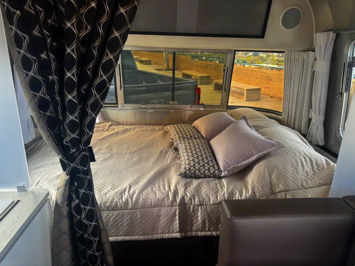Cozy rv rental interior with a quilted bed, stylish patterned curtains, and scenic outdoor views from the large windows.. Airstream 22' Airstream Caravel 2025