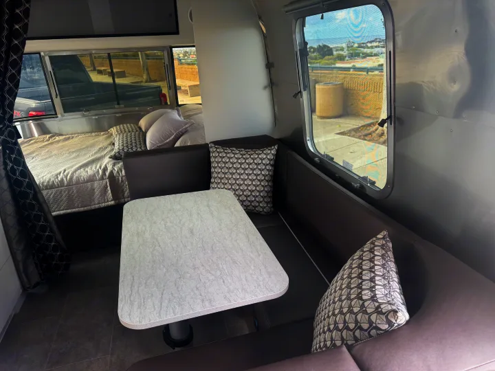 Bright RV rental interior: view of bed with pillows, textured dining table, and gray bench seating.. Airstream 22' Airstream Caravel 2025