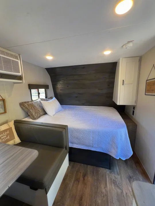 Modern RV rental's bedroom & dining area, featuring a bed with white quilt, AC, and a cozy bench with a table.. Keystone RV Hideout 2021