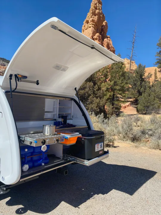 White teardrop trailer RV rental with its open rear hatch revealing a slide-out outdoor kitchen with cooktop, set against red rocks.. Bean Bean Stock 2021