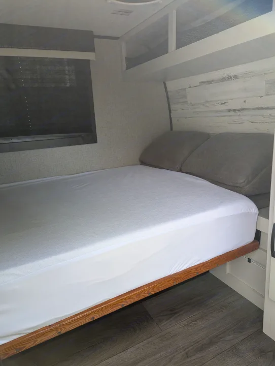Modern RV rental bedroom features a fresh white bed, grey pillows, and stylish wood-look decor. Window with blinds.. Jayco Jay Feather Micro 12SRK 2021