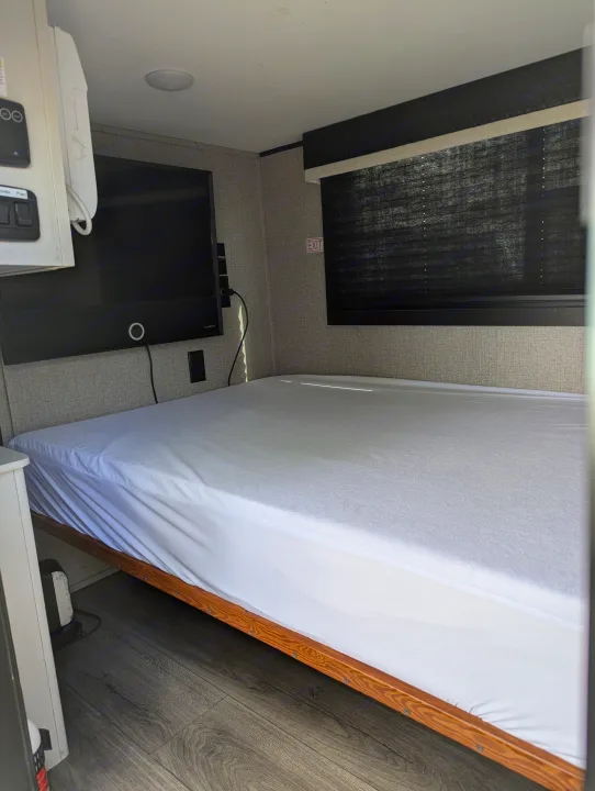Compact RV rental bedroom with white bed, wall-mounted TV, and window blinds, ready for travel.. Jayco Jay Feather Micro 12SRK 2021