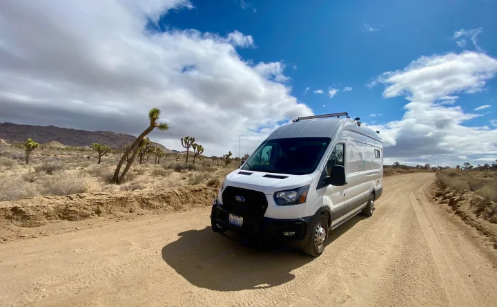 White RV rental van on a desert dirt road, surrounded by Joshua trees and mountains under a cloudy sky.. Ford Transit Transit 2022