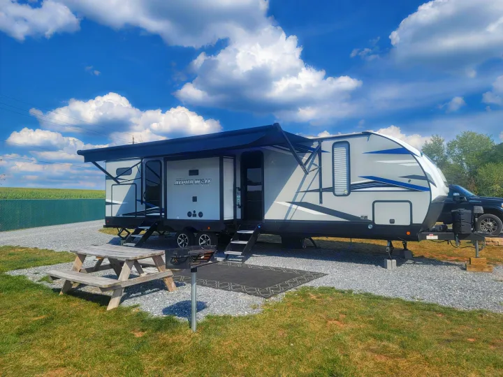 Serene rv rental campsite featuring a white & blue Alpha Wolf travel trailer, extended awning, picnic table, and grill.. Forest River Cherokee Alpha Wolf 2021