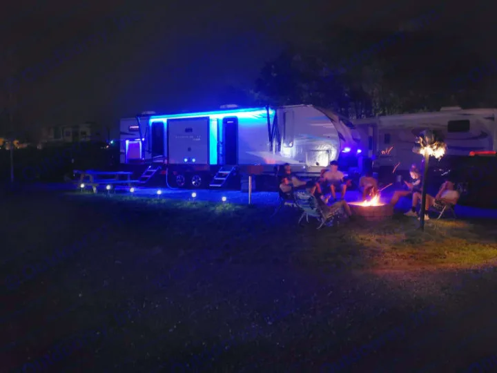 Nighttime RV rental scene: bright blue lights illuminate the camper and ground, while friends enjoy a warm campfire gathering.. Forest River Cherokee Alpha Wolf 2021