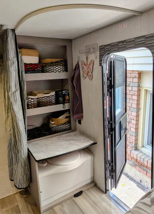 RV rental interior: organized storage shelves, compact toilet/counter, and an open exit door revealing the outdoors.. Coachmen Viking 1200ROK 2025
