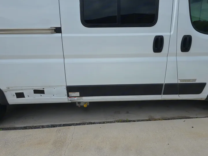 Side view of a white Winnebago RV rental, showing sliding door, windows, and external utility connections.. Winnebago Solis 2023
