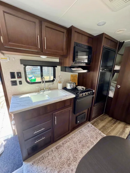 Inside an RV rental: dark wood kitchen with white sink, stove, microwave, fridge, and adjacent bunk beds for comfortable travel.. Jayco Jay Flight SLX 2019