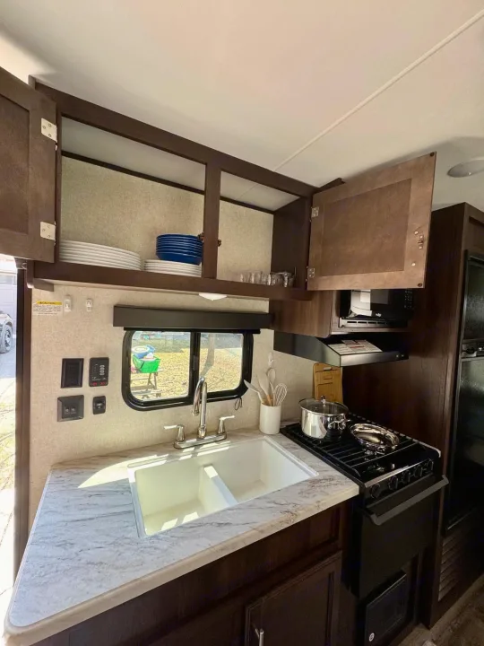 Cozy RV rental kitchen with a double sink, gas stove, microwave, and overhead cabinets stocked with dishes.. Jayco Jay Flight SLX 2019