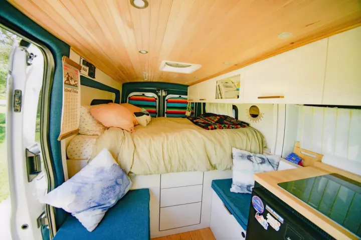 Inside a cozy RV rental: a comfortable bed at the back, wood-paneled ceiling, storage, and a compact kitchen area.. Ford Transit 2019