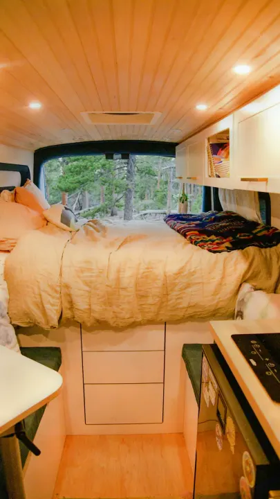 RV rental camper van interior. Features cozy bed looking out to forest, kitchenette, and wood-paneled ceiling.. Ford Transit 2019