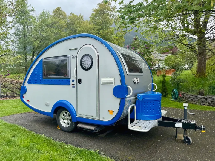 A blue & grey teardrop RV rental camper parked on asphalt, surrounded by green trees and grass, ready for travel.. Pleasant Valley/NuCamp T@B 2016