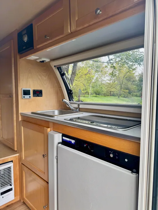 RV rental interior: compact kitchen with sink, stove, fridge, and control panels. Window view shows rain & trees.. Pleasant Valley/NuCamp T@B 2016