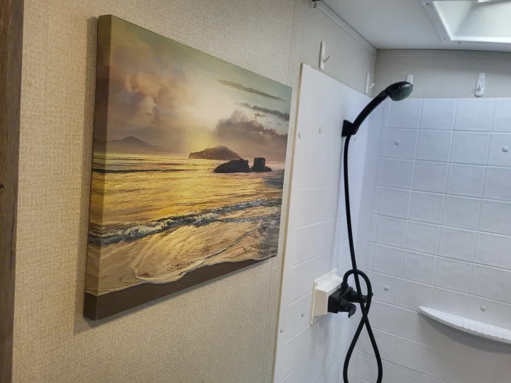 This RV rental shower features a serene beach sunset artwork on the wall, complementing the white shower stall with a black shower head.. Keystone Outback 2021