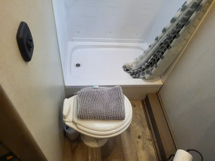 Inside an RV rental bathroom: a white shower stall with a polka-dot curtain and a toilet covered by a grey mat, on wood-look flooring.. Keystone Outback 2021