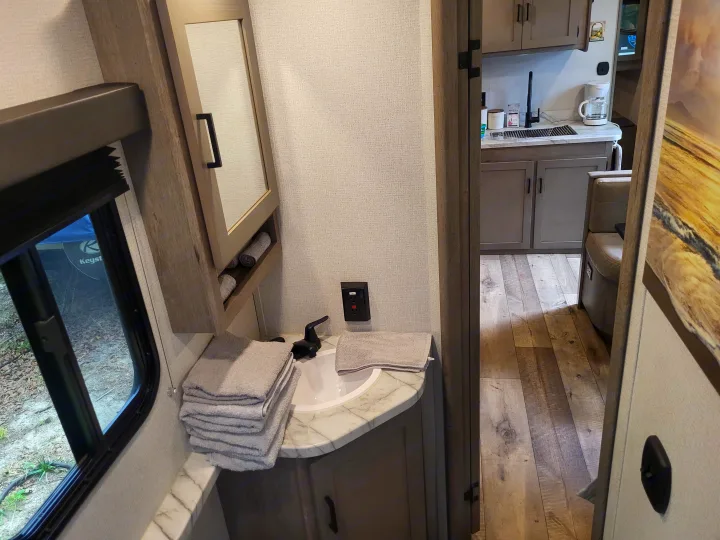 Compact RV rental bathroom sink and mirror, with a peek into the kitchen and living area, featuring wooden flooring.. Keystone Outback 2021