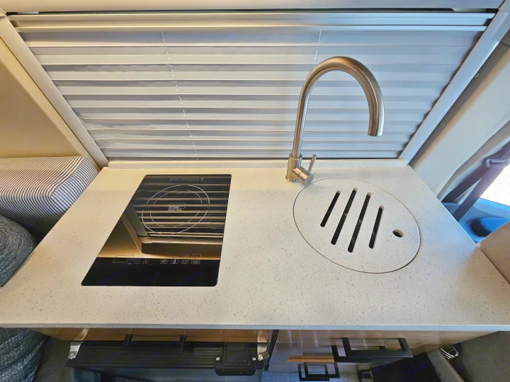 Compact RV rental kitchen featuring a modern induction cooktop, sink, and curved faucet. Pleated blinds overhead.. Forest River Coachmen Beyond 2023