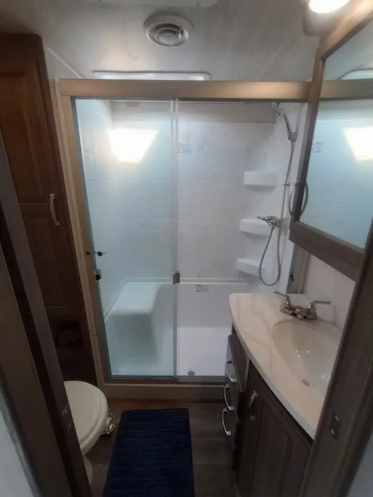 RV rental bathroom features a glass-enclosed shower with a seat, white toilet, and dark wood vanity with a sink.. Keystone RV Cougar 2022