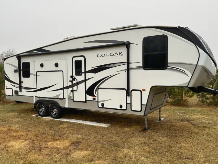 Modern white and black Cougar fifth-wheel RV rental. A large recreational vehicle parked on dry grass under an overcast sky.. Keystone RV Cougar 2022