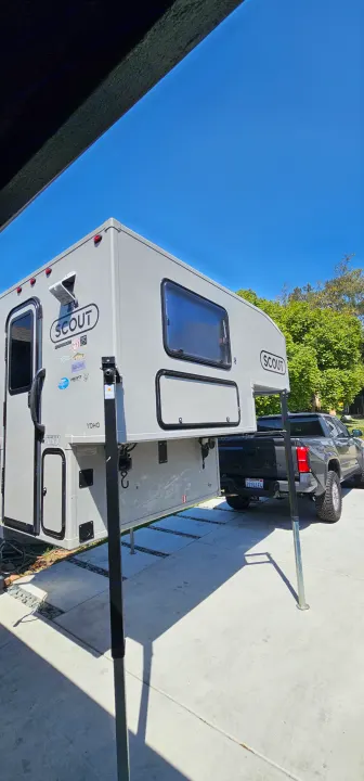 Light gray Scout Yoho truck camper on jacks next to a dark pickup truck on concrete. Perfect for RV rental outdoor adventures.. Toyota Tacoma 2025