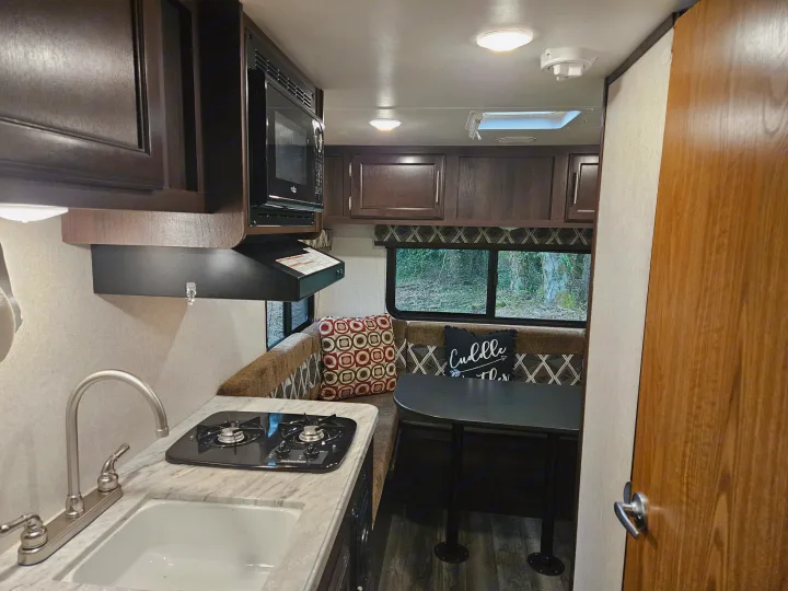 RV rental interior: a compact kitchen with sink, stove, & microwave, adjacent to a dining booth. Windows show outdoor trees.. Jayco Jay Flight SLX 2019