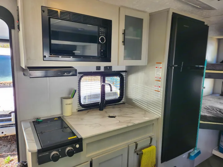 Modern RV rental kitchen features: microwave, stovetop, sink, and refrigerator. Light wood cabinets and a marble-look counter.. Forest River Wildwood FSX 2022