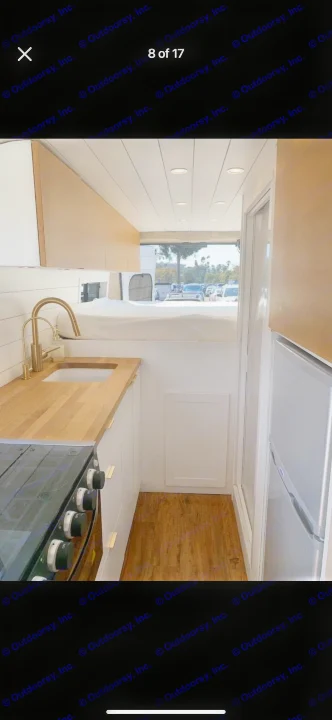 This RV rental interior features a compact kitchen with a sink, stove, and a bed overhead, offering cozy mobile living.. Custom Ram Promaster 2021