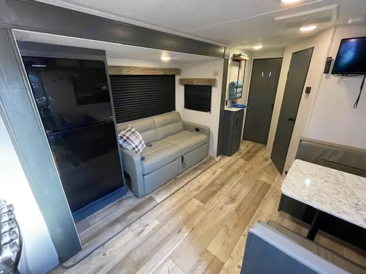 Inside a modern RV rental: gray sofa, black fridge, dinette, and wood-look floor in living area.. Dutchmen 23RKCWE 2024