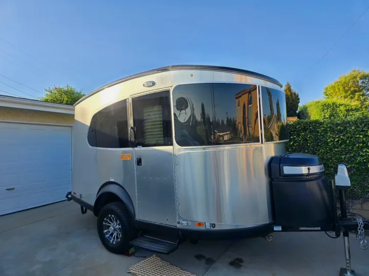 Sleek silver Airstream Basecamp-like travel trailer for RV rental, parked in a sunny driveway. Large windows reflect buildings & trees.. Airstream Basecamp 2021