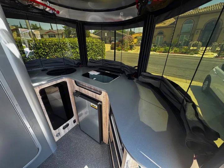 Modern RV rental interior with compact kitchen, fridge, and panoramic windows overlooking a sunny street scene.. Airstream Basecamp 2021