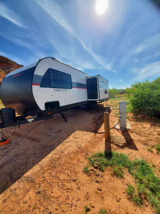 This RV rental is perfectly set up at a sunny desert campsite, featuring a travel trailer with hookups on reddish earth.. Forest River Salem Platinum 2024