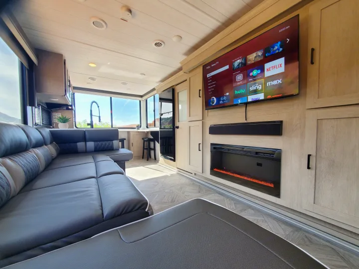 Bright rv rental living space with L-shaped sofa, large TV, electric fireplace, and kitchen area with scenic views.. Forest River Salem Platinum 2024