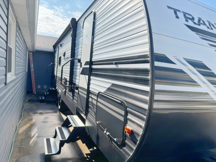 Sunny side view of a modern Transcend travel trailer parked beside a house, showing deployed steps. Great RV rental option!. Grand Design Transcend Xplor 2020